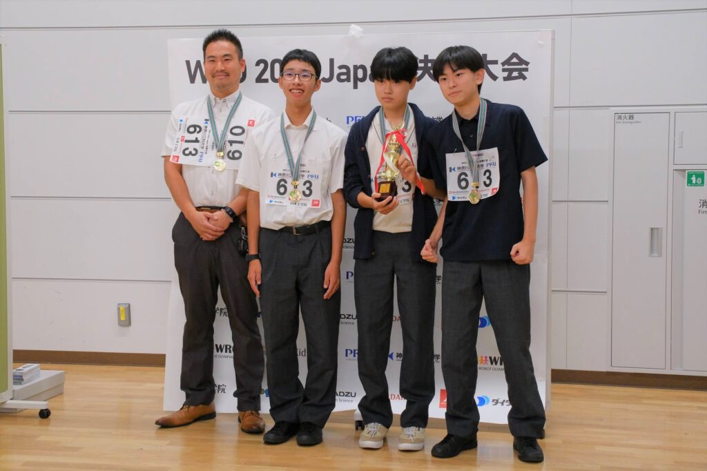 WRO 2023 Japan 決勝大会in Tokyoで「優勝」の記念撮影。左から、大澤教諭、「meiden1年チーム」の堀田くん、猪俣くん、楠本くん